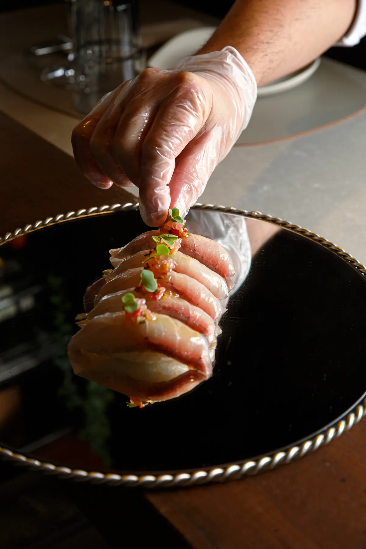 Una mano con guante coloca un nigiri sobre el plato de espejo negro